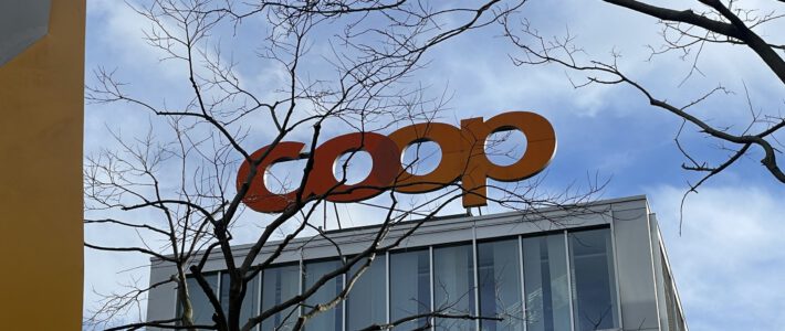 Ein Logo von Coop am Hauptsitz in Basel hinter Baumzweigen