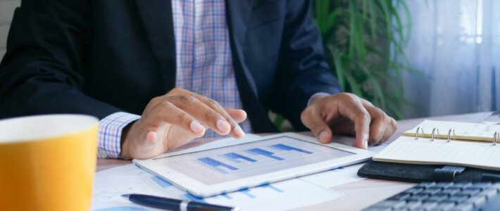 Ein Mann im Sakko vor einem Tablett mit Grafiken und einer Kaffeetasse sowie einem Taschenrechner