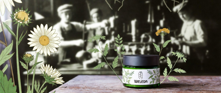 Blumen und ein Weleda-Produkt auf einem Tisch vor verschwommenen Arbeitern im Hintergrund