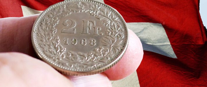 Eine 2-Franken-Münze auf zwei Fingern vor einer Schweizerflagge