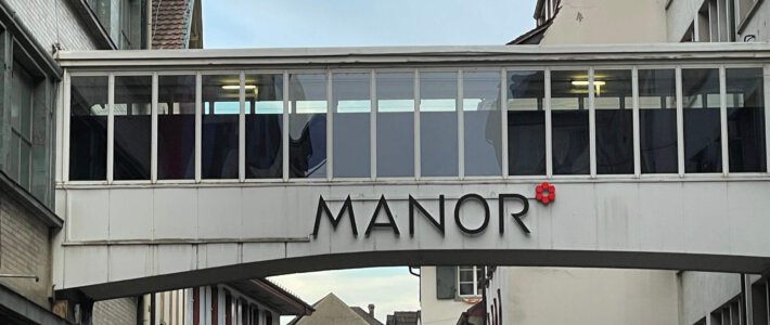 Trifft Manor eine Schuld für Basler Legionellenausbruch? Logo von Manor an einem Gebäudeübergang in Basel
